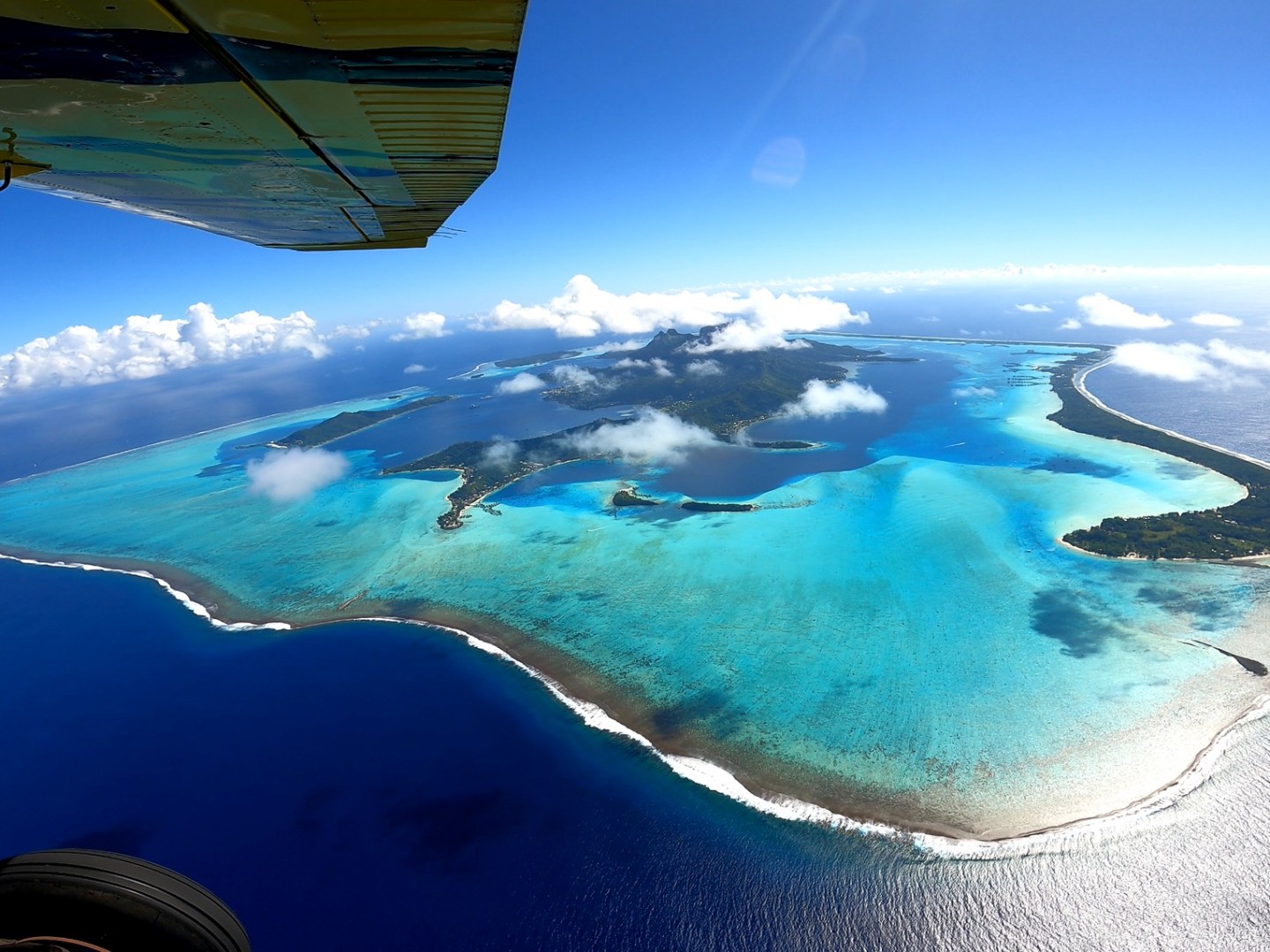 Aerial view of a tropical island with turquoise lagoon under a bright blue sky, partially obscured by airplane wing.