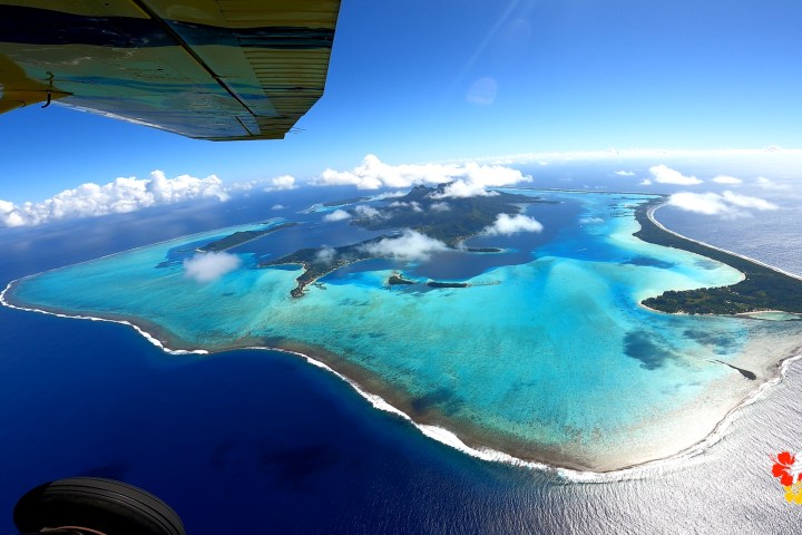 Aerial view of a tropical island with turquoise lagoon under a bright blue sky, partially obscured by airplane wing.