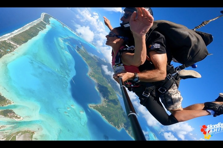 Two people tandem skydiving over turquoise waters and lush islands.