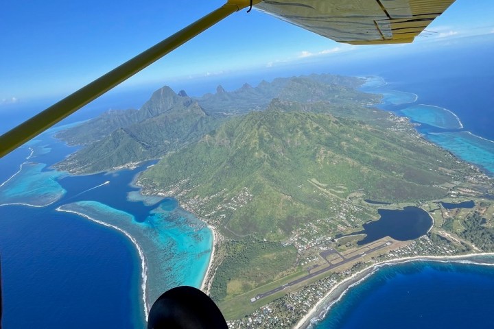 Aerial view of a tropical island with an airplane wing overhead and award logo in corner.