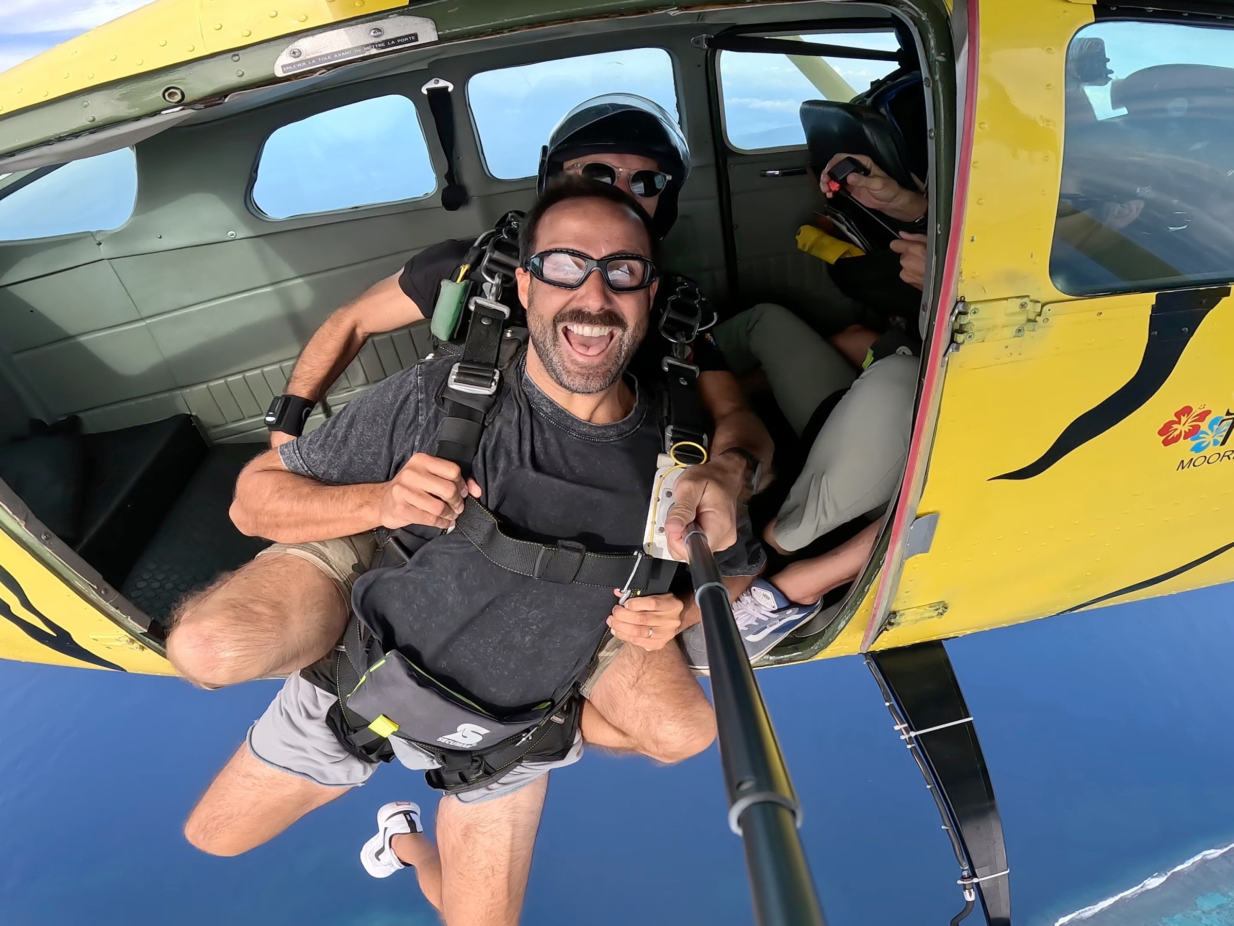 Man with selfie stick skydiving tandem from yellow plane with open door, ocean below.