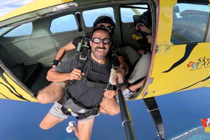 Man with selfie stick skydiving tandem from yellow plane with open door, ocean below.