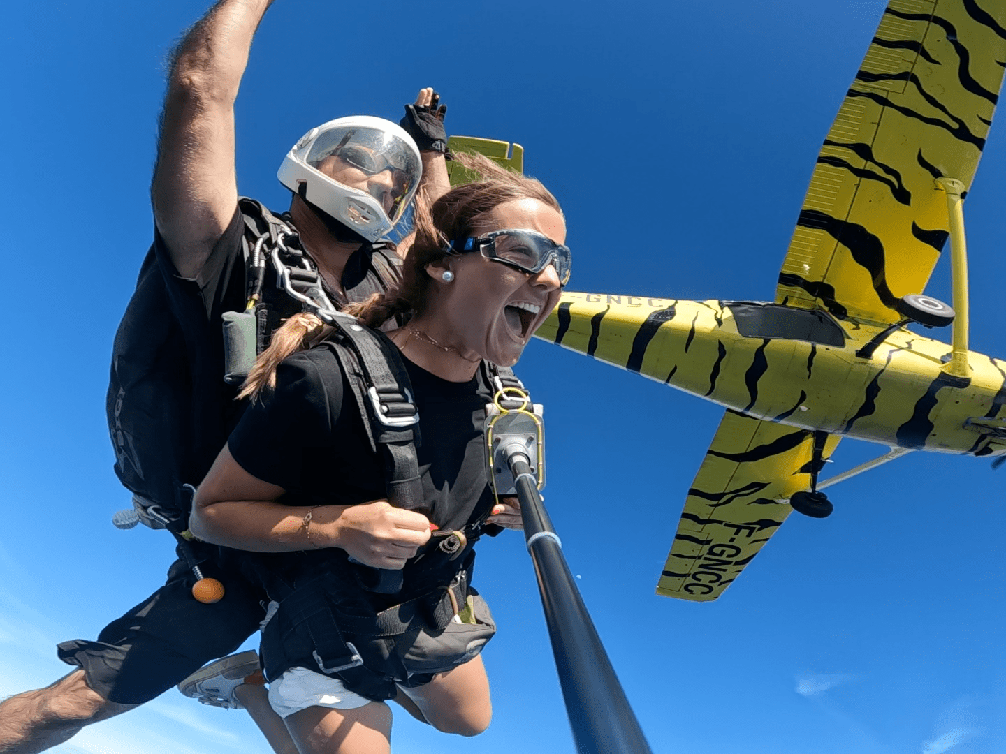Two people tandem skydiving near a yellow tiger-striped airplane.