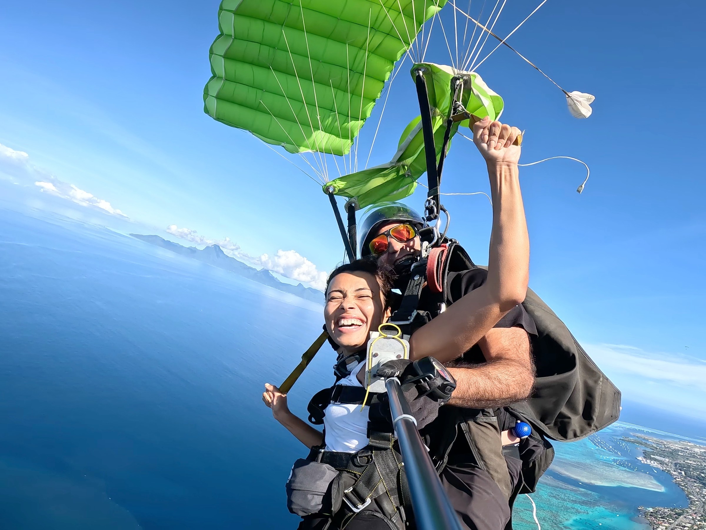 Two people tandem skydiving over the ocean with a green parachute, one smiling and holding a selfie stick.