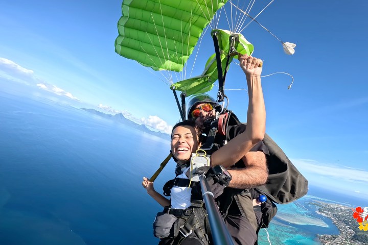 Two people tandem skydiving over the ocean with a green parachute, one smiling and holding a selfie stick.