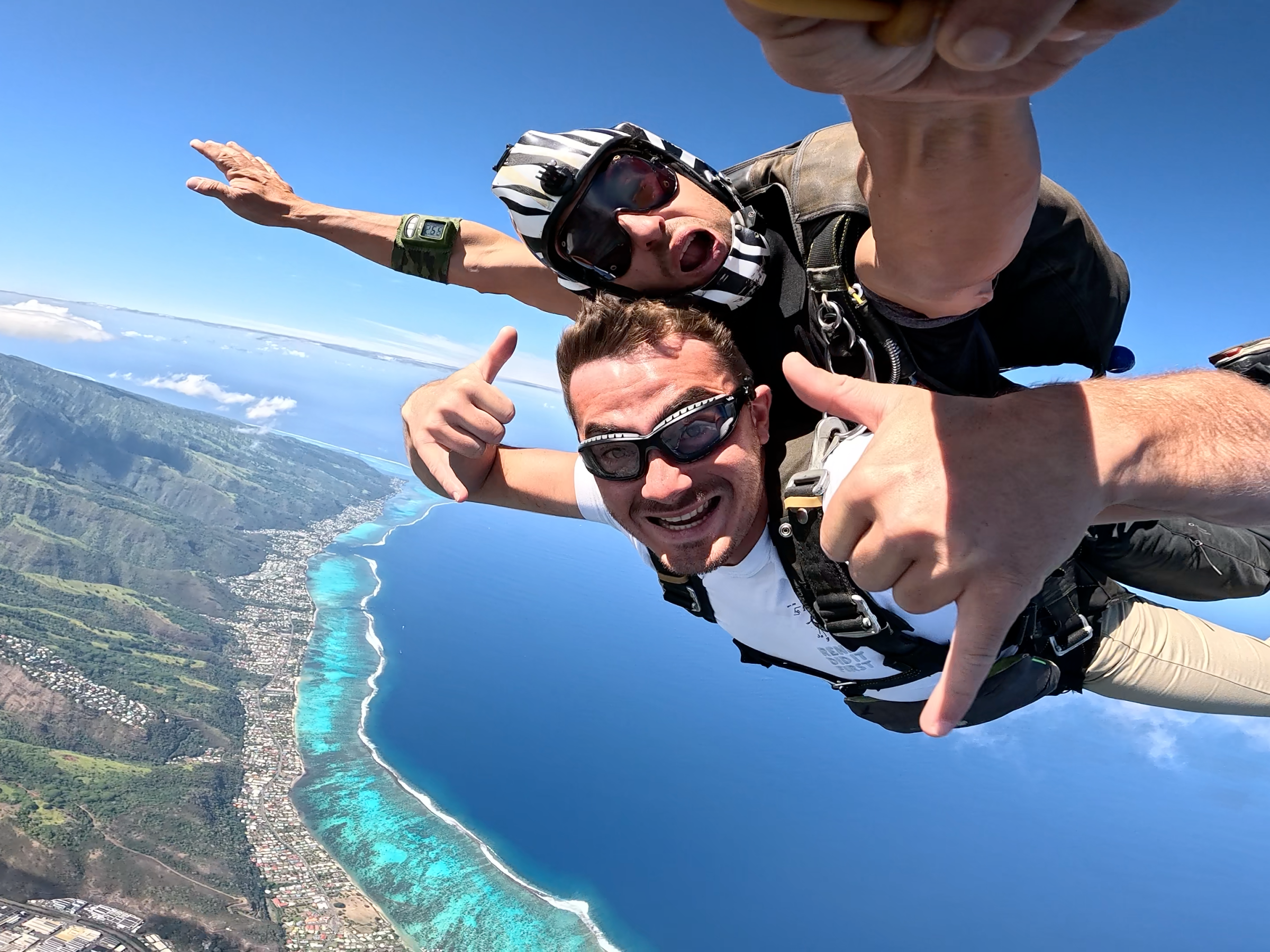 Two people tandem skydiving over a coastal landscape, smiling and giving thumbs up.