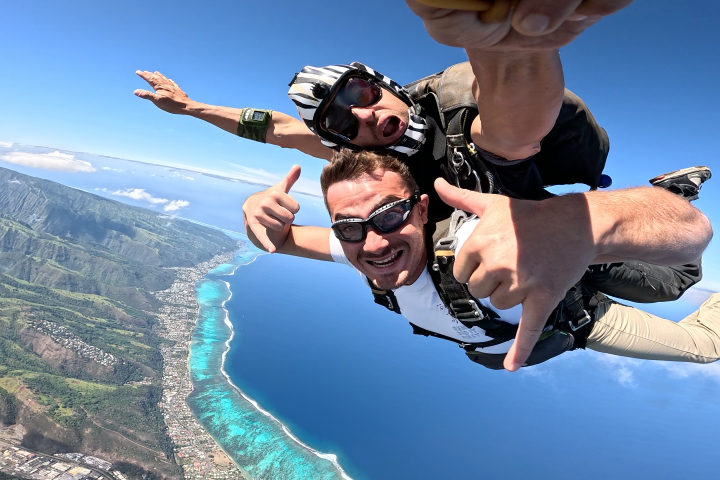 Two people tandem skydiving over a coastal landscape, smiling and giving thumbs up.