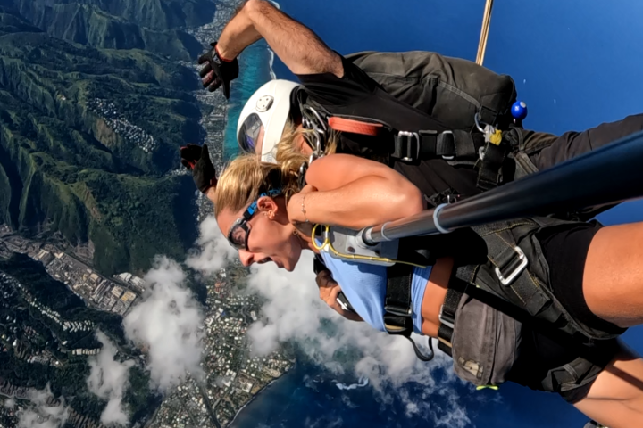Two people tandem skydiving over Tahiti with ocean and mountains in view, Skydive Tahiti logo visible.