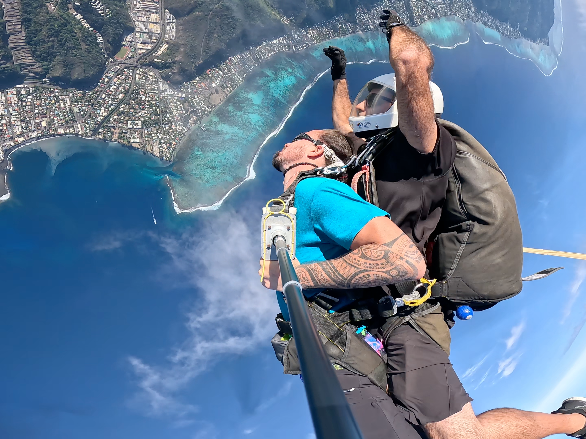 Tandem skydivers free-fall above an island coastline with clear blue water and green land.