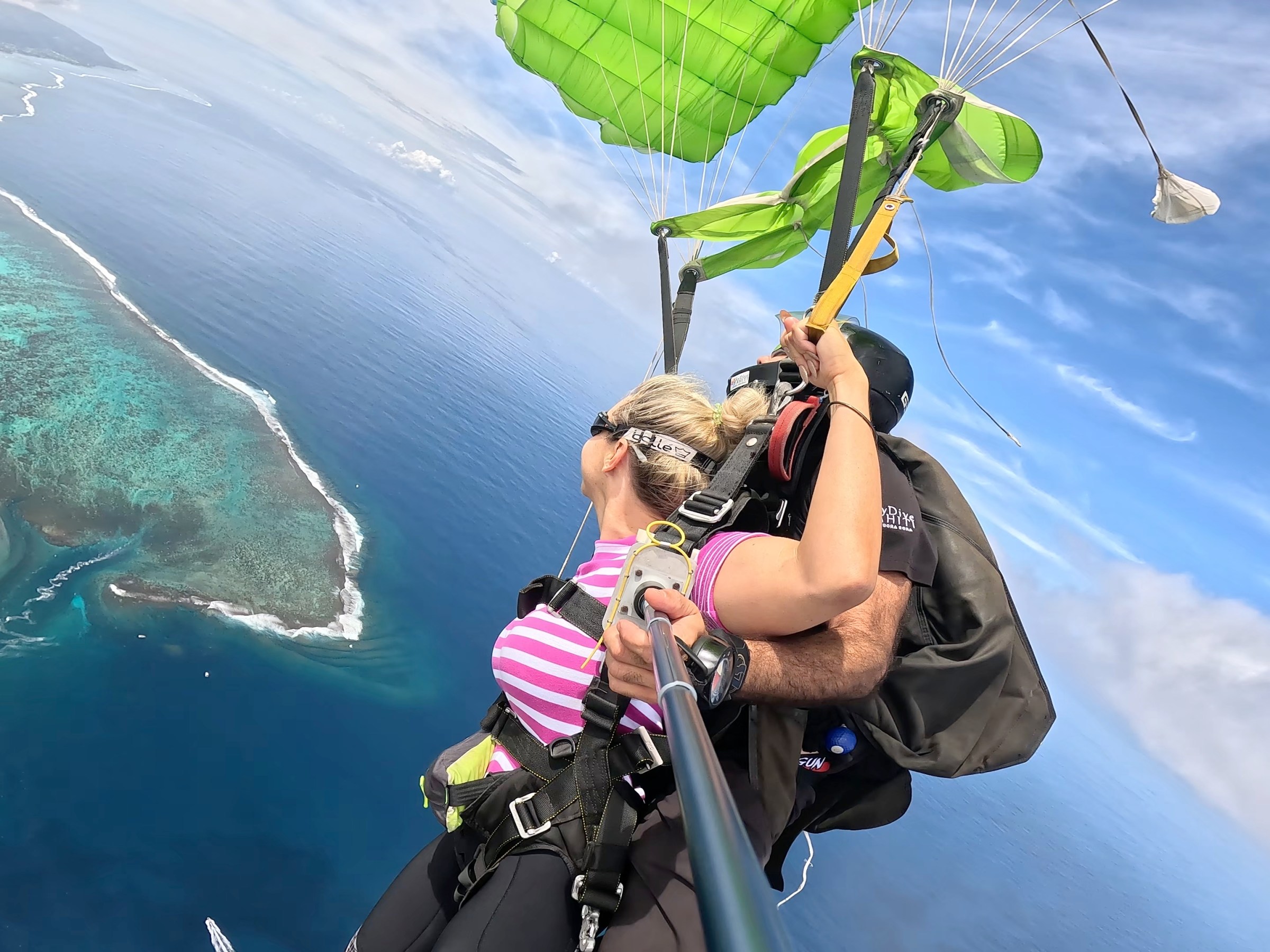 Two people tandem skydiving over a coastal area with a green parachute.