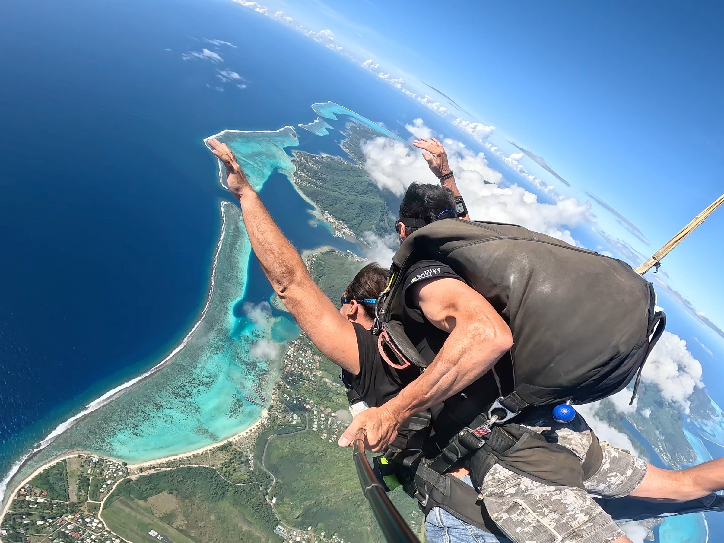 Tandem skydive over turquoise ocean and lush islands, parachute deployed, sunny skies.